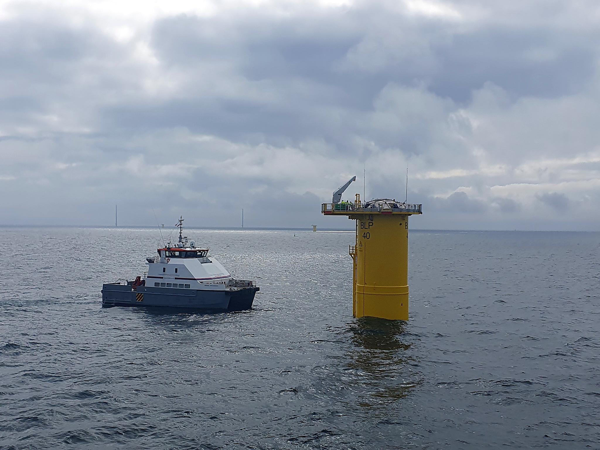 A crew transfer vessel working alongside a yellow offshore wind foundation platform beneath a cloudy sky.