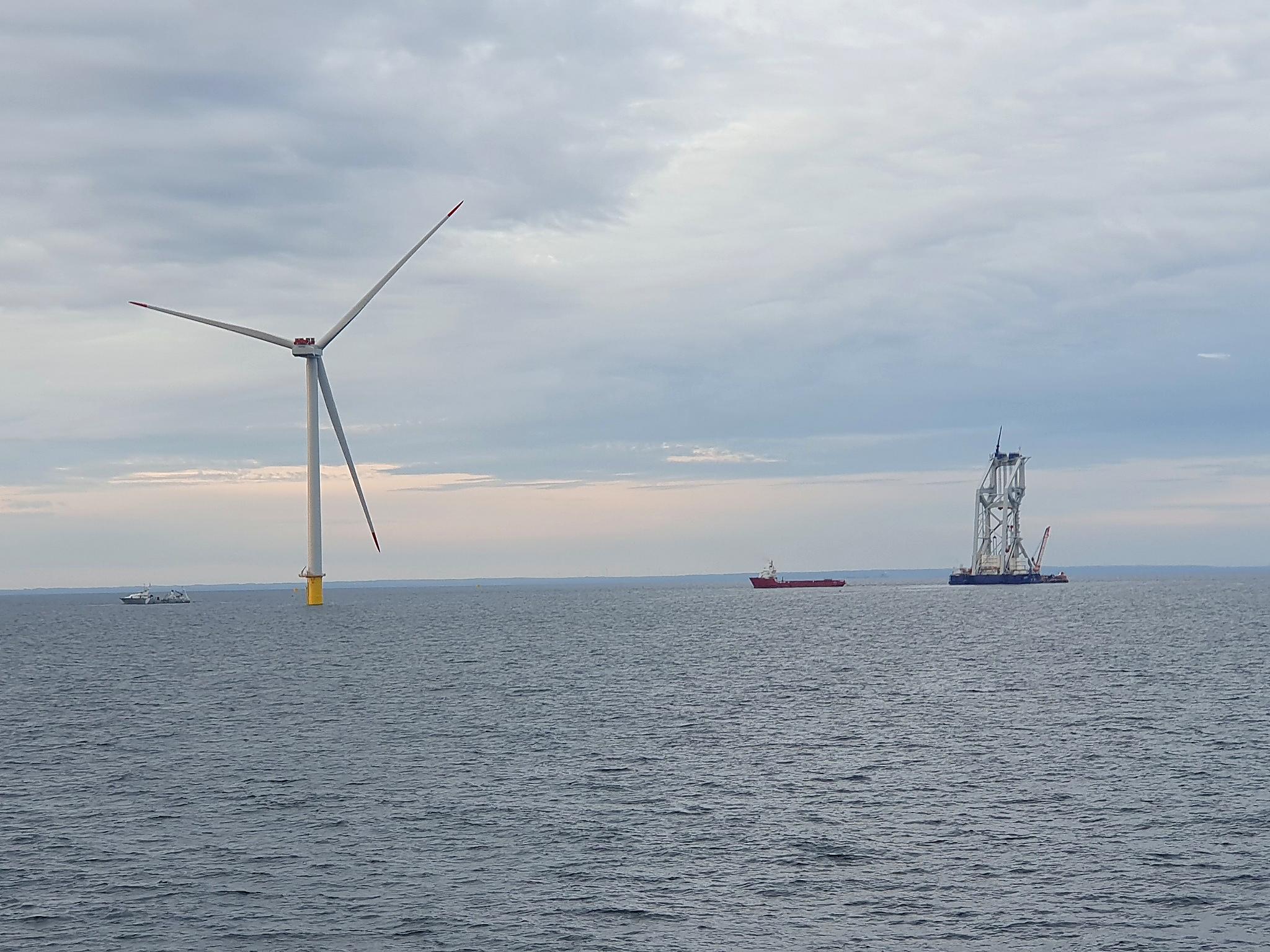 An offshore wind turbine rising from calm water, with a red cargo ship and a jack-up installation vessel on the horizon.