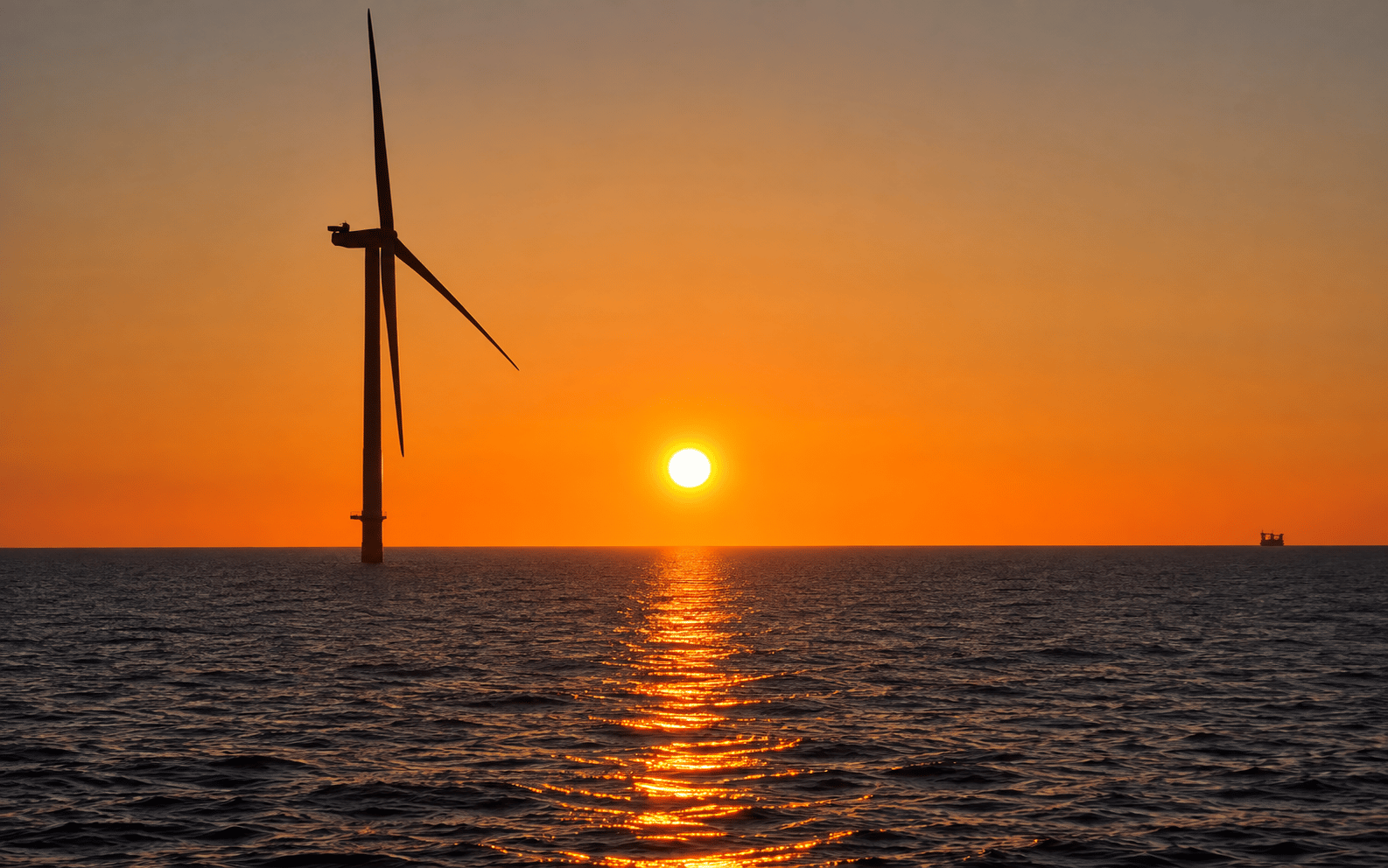 An offshore wind turbine silhouetted against an orange sunset, with the sun reflecting a glitter path across the sea.